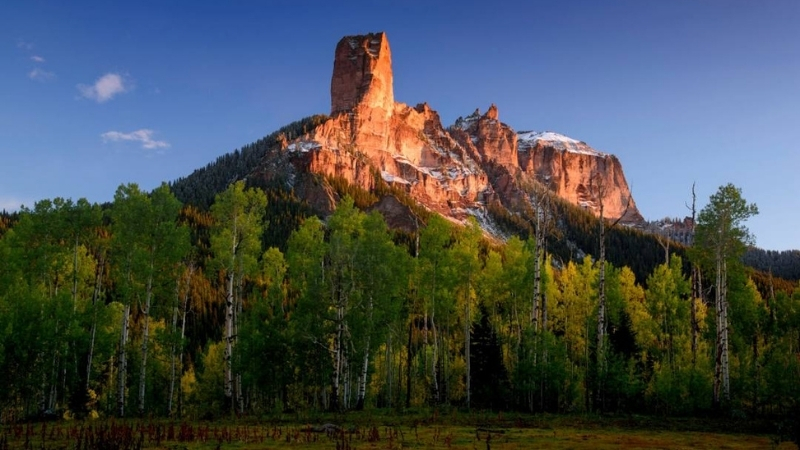 Towering rock formation bathed in warm sunlight with green aspen trees in the foreground and a dusting of snow on the mountain peak.